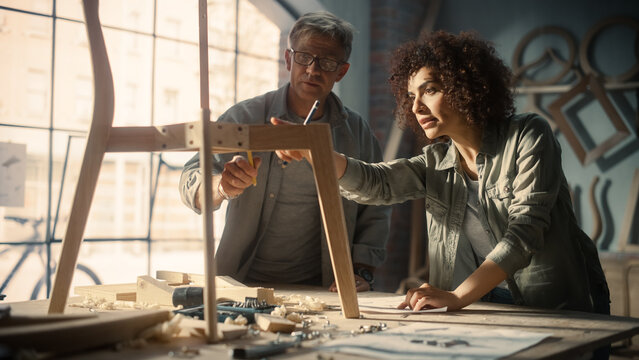 Fototapete - Portrait of Two Small Business Owners Working Together on Creating a New Wooden Dining Table Chair Design. Adult Man and Young Beautiful Female Looking at a Blueprint and Discussing the Work Process.