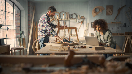 Two Carpenters Working Together in a Woodwork Workshop. Multicultural Man and Female Colleagues Looking at a Blueprint on Laptop Computer, Creating a New Chair Design.