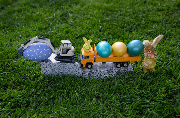 Wall Mural - models of toy gray excavator and yellow dump truck stand on granite stone in grass along with bright colorful Easter eggs. Easter spring holiday concept for congratulations from construction companies