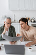 © LIGHTFIELD STUDIOS - young man pointing with hand near thoughtful dad holding folder with documents while sitting at laptop