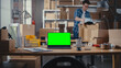 © Gorodenkoff - Desktop Computer Monitor Standing on a Table with a Green Screen Chromakey Mock Up Display. Small Business Warehouse with Worker Walking in the Background. Desk with Cardboard Boxes