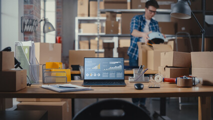 Laptop Computer Monitor Standing on a Table with a Trend Analysis Charts Display. Small Business Storage Room with Worker Walking in the Background. Desk with Cardboard Boxes.