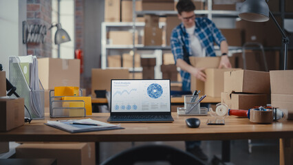 Wall Mural - Desktop Computer Monitor Standing on a Table with a Trend Analysis Charts Display. Small Business Warehouse with Worker Walking in the Background. Desk with Cardboard Boxes