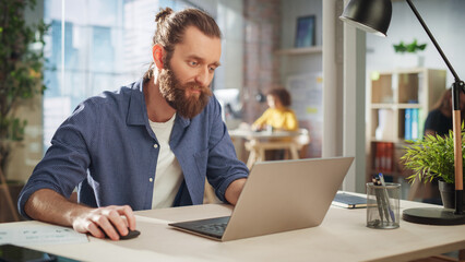 Wall Mural - Close Up Portrait of a Handsome Long-Haired Bearded Manager Sitting at a Desk in Creative Office. Stylish Man Using Laptop Computer in Marketing Company. Writing Inspiring Startup Business Ideas.