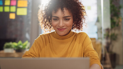 Poster - Close Up Portrait of a Happy Middle Eastern Manager Sitting at a Desk in Creative Office. Young Stylish Female with Curly Hair Using Laptop Computer in Marketing Agency.