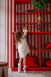 Cute little curly-haired girl in dress on tiptoe reaches for xmas bell on shelf