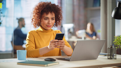 Aufkleber - Beautiful Middle Eastern Manager Sitting at a Desk in Creative Office, Using Smartphone. Young Stylish Female Sending Message, Browsing Internet. Colleagues Working in the Background.