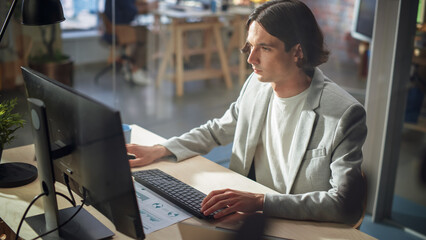 Poster - Stylish Long-Haired Manager Wearing a Gray Jacket, Working at a Desk in Creative Office. Young Handsome Man Using Desktop Computer in Marketing Agency. Colleagues Working in the Background.