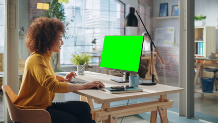 Poster - Portrait of an Excited Multiethnic Specialist Working in Creative Agency. Stylish Expressive Female in Yellow Jumper Using Desktop Computer with Green Screen Mock Up Display.