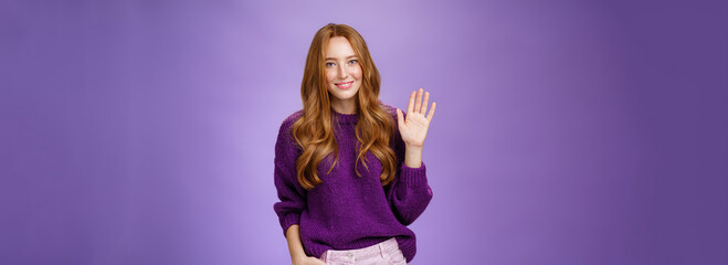 Wall Mural - Shy and cute redhead female newbie greeting new team smiling friendly and waving raised palm in hello or hi gesture, saying nice to meet you as getting know new member over violet wall