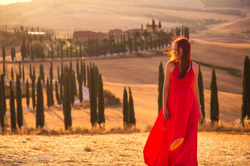 A girl at sunset in a red dress on a field in Italian Tuscany. Val d'Orcia. Beautiful landscape scenery at sunset of Tuscany in Italy