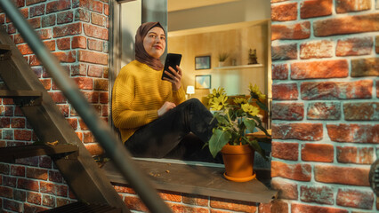Aufkleber - Portrait of a Young Arab Woman Using a Smartphone while Sitting on a Window Sill in a Multi-Storey Brick House. Multiethnic Female Connecting with Friends Online. Shot from Outside the House.