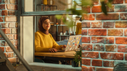 Fototapete - Young Muslim Female Student Using Laptop at Home. Attending Remote Education During the Day. Working on Computer, Writing an Essay Assignment. Arc Shot from Outside the Window.