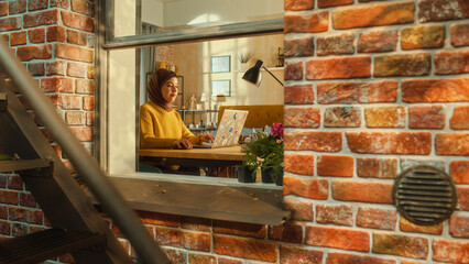 Poster - Young Muslim Female Using Laptop at Home. Doing Remote Work During the Day. Beautiful Girl in a Hijab Sitting at a Table, Working On Computer, Brainstorming Creative Project. Outside the Window Shot.