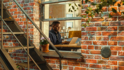 Wall Mural - Young Handsome Male Using Laptop Computer, Sitting in Living Room in Apartment. African American Businessman Answering Emails, Writing a Business Plan from Home. Inside Apartment Window View.