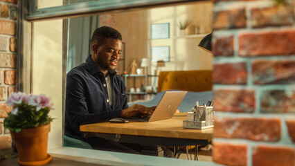 Poster - Young Handsome Male Using Laptop Computer, Sitting in Living Room in Apartment. African American Businessman Answering Emails, Writing a Business Plan from Home. Inside Apartment Window View.