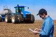 © Designpics - Farmer using a tablet while standing on a farm field and a tractor and equipment seeds the field; Alberta, Canada