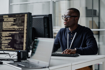 African American young developer in eyeglasses concentrating on his online work on computer sitting at workplace