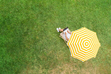 Woman In A White Dress Sitting On Deck Chair Under Yellow Umbrella With Laptop On The Green Grass Sunbathes At Summer Day. Top View, Drone, Aerial View.