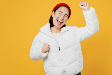 Wall Mural - Young overjoyed woman of Asian ethnicity wear white padded windbreaker jacket red hat do winner gesture celebrate clenching fists isolated on plain yellow background studio People lifestyle concept.