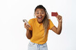 Happy young black woman showing her credit card after using mobile phone app, laughing with joyful face expression, white background
