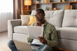 © Prostock-studio - Pensive serious adult black man in glasses uses computer, works with documents, sits on floor in living room