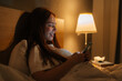 Side view of cheerful redhead young woman using smartphone, looking on screen, typing online message on social media, lying on bed late at night, bedside lamp lighting with warm yellow light.