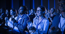 Young Woman Sitting In A Crowded Audience At A Business Conference. Female Attendee Cheering And Clapping After A Motivational Keynote Speech. Auditorium With Young Successful Businesspeople.