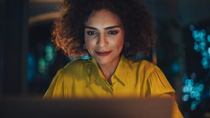 Wall Mural - Close Up Portrait of a Happy Middle Eastern Manager Sitting at a Desk in Creative Office. Young Stylish Female with Curly Hair Using Laptop Computer in Marketing Agency in the Evening.