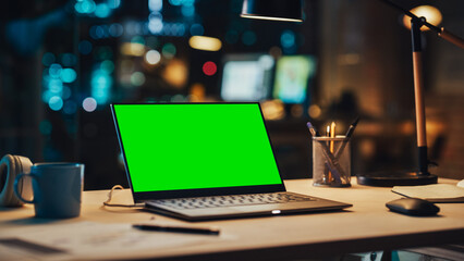 Poster - Laptop Standing on a Desk with a Green Screen Chromakey Mock Up Display. Table with Computer, Coffee Mug, Headphones and Notebook in Creative Office at Night.