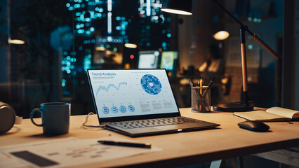 Poster - Laptop Standing on a Desk with Sales Statistics and Data Chart Displayed on Screen. Table with Computer, Coffee Mug, Headphones and Notebook in Creative Office at Night.