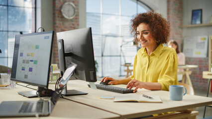 Poster - Portrait of Biracial Creative Young Woman Working on a Computer in Bright Busy Office. Female Recruiter Smiling Happily When Receiving Positive Feedback. Employee Working in Background.