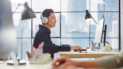 Poster - Young Asian Man Working on a Computer and Listening Through Headphones in a Modern Office. Male Client Advisor Answering Requests. Big Window with a City View in the Background.