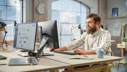 Fototapete - Portrait of Creative Bearded White Man with Glasses Smiling While Using Computer in Bright Spacious Office. Male Team Manager Giving Feedback to colleagues. People Working in Background.
