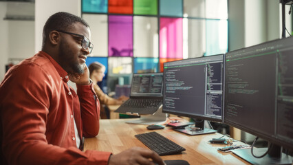 Wall Mural - Black Man Writing Lines of Code On Desktop PC With Two Monitors and a Laptop in Stylish Office. Professional Male Developer Programming Artificial Intelligence Software for Start-Up Company.