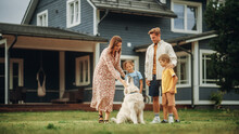 Portrait Of A Cheerful Family Couple With Kids, Playing And Petting A Beautiful White Golden Retriever Dog. Happy Successful People Standing On A Lawn In Their Front Yard In Front Of The House.