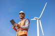 Engineer India man working with tablet at windmill farm Generating electricity clean energy. Wind turbine farm generator by alternative green energy. Asian engineer checking control electric power