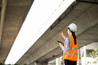 © Chanakon - Asian engineer worker woman or architect looking construction with tablet wear white safety helmet in construction site. Standing at highway concrete road site. Progress planning of highway bridge.