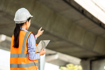 Wall Mural - Asian engineer worker woman or architect looking construction with tablet wear white safety helmet in construction site. Standing at highway concrete road site. Progress planning of highway bridge.