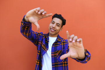 Wall Mural - Young man of hispanic heritage wearing colorful flannel shirt showing a camera frame gesture over orange wall. Copy space for text, close up, background.