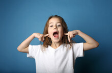 A Little Girl Screams With Her Mouth Wide Open And Her Ears Closed Against A Blue Background