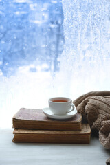 Wall Mural - tea cup, book, sweater and winter frozen window abstract background. cozy mood, home comfort in snowy cold weather. festive winter season. Christmas, New Year holidays