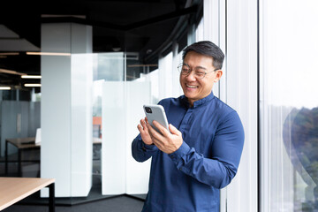 Asian businessman inside the office in a casual shirt and glasses is using the phone, the boss is holding a smartphone in his hands reading news and typing a message smiling.