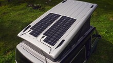 Orbit Drone Shot Of Silver Camper Van With Two Solar Panels On Opened Pop Top Roof Tent. Car Parked On Grass In Ancient Forest.
