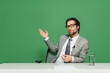 © LIGHTFIELD STUDIOS - broadcaster in eyeglasses and grey suit sitting at desk and pointing with hand isolated on green