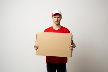 Canvas Print - Portrait of a young bearded delivery man in red uniform holding blank cardboard, percel isolated on white background.