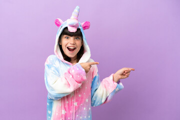 Poster - Little kid wearing a unicorn pajama isolated on purple background surprised and pointing side