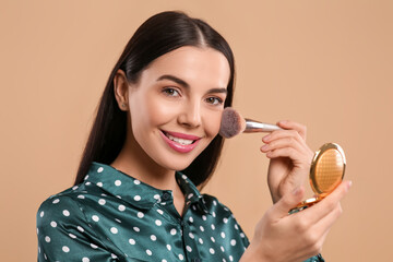 Happy woman with cosmetic pocket mirror applying makeup on light brown background