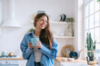 © Iona - Happy caucasian female with long loose hair in jeans and blue shirt toothy smiles holds cup of tea looks aside happily enjoys weekend at cozy home. Pretty hispanic woman laughs against blurry kitchen.