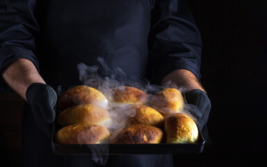 Chef holds a baking sheet with freshlybaked buns or pies. Black space for menu or recipe.